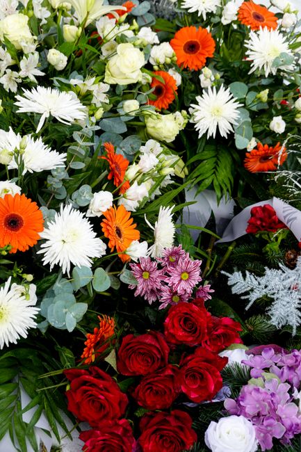 A vibrant floral arrangement featuring a mix of flowers, including bright orange gerbera daisies, white asters, soft white roses, pink daisy-like flowers, and deep red roses. The bouquet is densely co