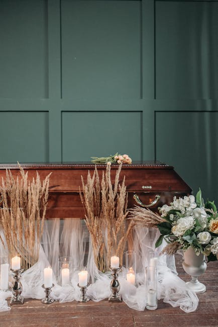 A collection of funeral floral arrangements displayed in front of a wooden casket with a dark wood finish, set against a green paneled wall. The arrangements include a large bouquet in a white vase wi