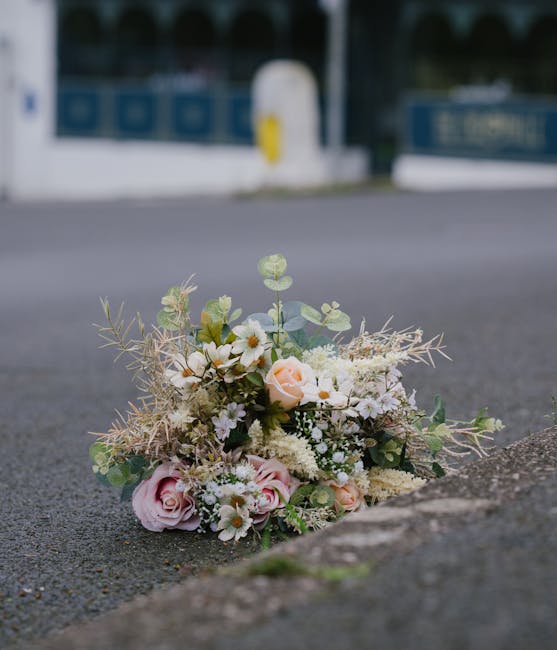 A delicate bouquet of fresh flowers rests on an asphalt surface near the curb, featuring soft pink roses, white daisies, and cream-colored blooms, complemented by greenery and dried foliage for textur
