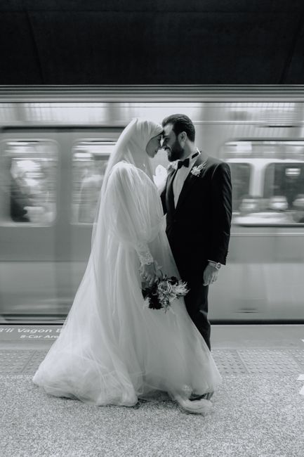 A bride and groom share an intimate moment on a train platform, with the bride dressed in a flowing white wedding gown and veil, holding a small bouquet of mixed flowers featuring roses, carnations, a