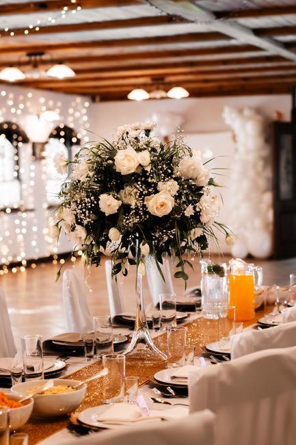 A tall floral arrangement featuring predominantly white roses and delicate baby's breath, accented with lush green foliage, is displayed in a clear glass vase. The bouquet is set atop a wooden dining