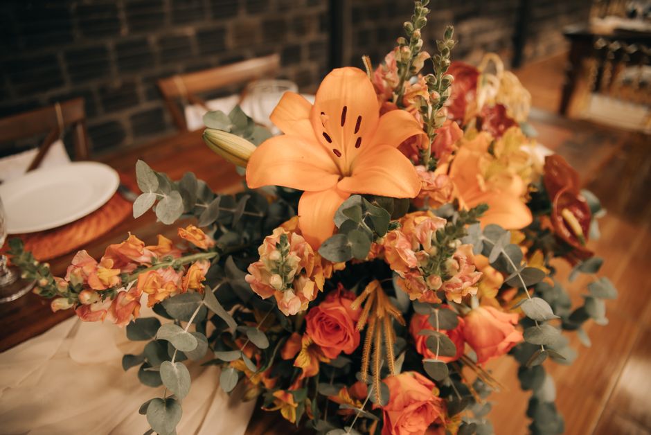 A floral arrangement featuring prominent orange lilies with wide, curved petals and dark stamens, complemented by peach and coral roses with tightly coiled petals, and clusters of pink and peach stock